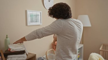 Genç İspanyol adam, beşikte rahat yatak odası ortamında, yumuşak ışıklandırma ve ahşap bir beşik ve lüks bir dekor gibi gerekli bebek eşyalarıyla çevrili, sıcaklık ve bakım taşıyan bir bebek beşiği taşıyor..