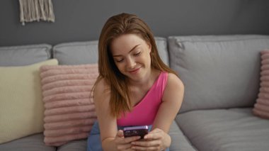 Kızıl saçlı, gülümseyen ve akıllı telefonuna bakan genç bir kadın şık bir oturma odasında rahat bir kanepede oturuyor.