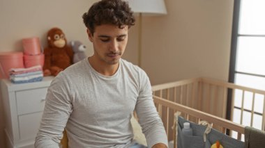 Genç adam rahat bir yatak odasında beşikte oturuyor, neşeli bir dekor, yumuşak ışıklandırma, ve ev ortamında huzurlu bir atmosfer, sakin bir aile anını yakalıyor..