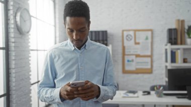 Bir ofis odasında raflar ve belgelerle çevrili akıllı bir telefonla duran genç adam, odaklanmış ve profesyonel bir çalışma ortamı olduğunu gösteriyor.