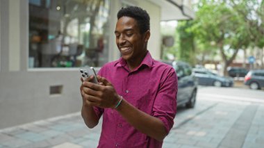 Şehirdeki şehir merkezinde akıllı telefon kullanırken gülümseyen genç bir adam. Arka planda arabalar ve ağaçlar görülebiliyor. Güneşli bir gün havası var..