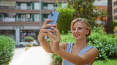 Şehir parkında selfie çeken bir kadın, kısa sarı saçlarıyla gülümsüyor, arka planda yeşillik ve şehir binalarıyla çevrili.