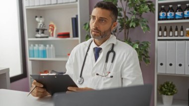 Modern klinik odasında beyaz önlük ve steteskop giyen yakışıklı doktor, profesyonel içerik içeriğinde not panosu ve kalem tutarken özenli ifadeler gösteriyor..