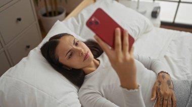 Genç bir kadın beyaz çarşaflı ve düşünceli bir ifadeyle modern bir yatak odasında akıllı telefon kullanarak yatakta yatıyor.