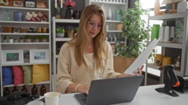 Ev dekorasyonu mağazasında dizüstü bilgisayarda çalışan sarışın bir kadın çeşitli dekoratif malzemelerle çevrili, iyi organize edilmiş, kapalı bir ortamda odaklanmış bir ifadeyle belgeleri inceliyor..