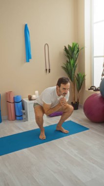 İyi ışıklandırılmış sağlık odasında yoga yapan adam egzersiz ekipmanları ve bitkilerle çevrili, dingin ve dengeli bir ev ortamını taşıyor..