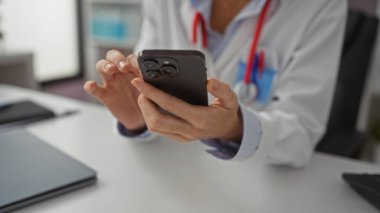 Klinikte beyaz önlüklü bir kadın masasında ellerini ve tıbbi ekipmanlarını gösteren, sağlık teknolojisi ve iletişim temasını taşıyan akıllı telefon kullanıyor..