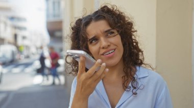 Şehir sokağında telefonla konuşan genç bir kadın ve gün boyunca arka planda binalar var.