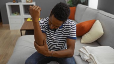 Oturma odasında dirseğini tutan genç bir adam modern bir dairede rahat bir koltukta otururken rahatsızlık göstererek günlük ev dekorasyonunu yansıtıyor..