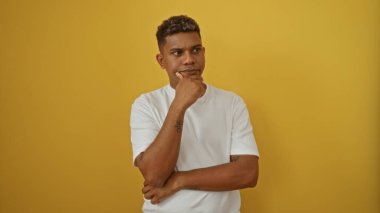 Düşünceli genç İspanyol adam, parlak sarı bir arka planda izole edilmiş, beyaz tişört içinde dalgın görünüyor.