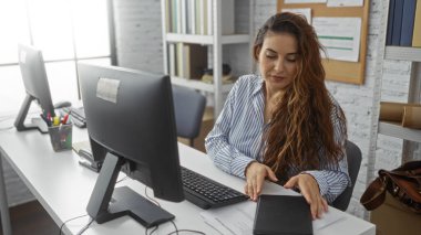 Bilgisayarlı bir ofiste çalışan genç İspanyol bir kadın, evrak işlerine odaklanıyor, modern kapalı bir çalışma alanı ve iş ortamı sunuyor..