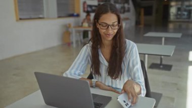 Ofisteki genç kadın bilgisayardaki video görüşmesi sırasında akıllı telefona bakıyor ve gülümsüyor.