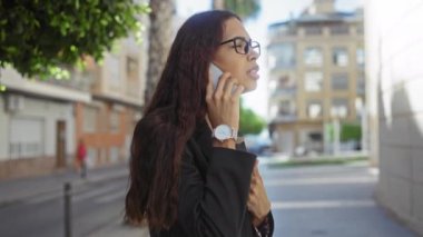 Gündüz vakti şehir sokağında yürürken akıllı telefondan konuşan genç bir kadın siyah ceket ve gözlük takıyor.