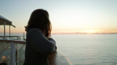 Gemi güvertesinde gün batımından hoşlanan bir kadın, gülümsüyor, okyanus manzarasının ortasında rahat, rahat gri süveter giyiyor, sakin bir atmosfer, canlı gökyüzüne karşı, açık havada seyahat deneyimi yakaladı..