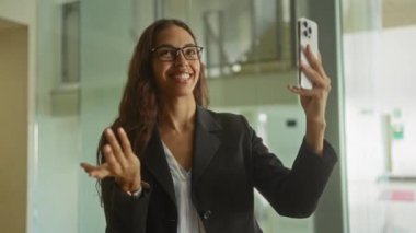 Modern ofis içinde akıllı telefondan konuşan bir kadın. Video görüşmesi sırasında gülümseyen ve el kol hareketi yapan.