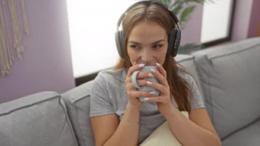 Kanepede oturan, kulaklıklı, içkisini yudumlayan, rahat, mor duvarları olan bir kadın..