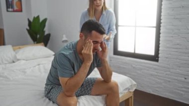 Kadın yatak odasında dikilirken, erkek yatakta oturuyor ve ev ortamında ilişki gerilimini gösteriyor..