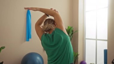 Spor salonunda esneyen, yeşil bluzlu, mavi spor topu ve duvarda asılı havlusu olan bir kadın.