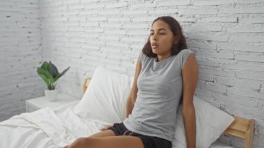 Beyaz tuğladan duvarları olan bir yatak odasında oturan, gri bir gömlek giyen, düşünceli ve düşünceli görünen bir kadın..
