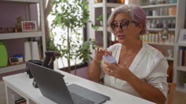 Bir kadın dizüstü bilgisayar ekranından konuşuyor ve çevrimiçi etkileşim sırasında meşgul ve heyecanlı görünen çeşitli eşyalarla dolu rahat bir odada ürünleri gösteriyor..