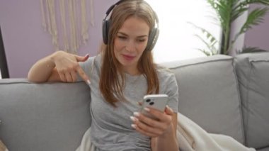 Akıllı telefondan bir video görüşmesi yapan, kulaklık takan ve günlük gri tişörtler giyen, rahat bir oturma odasında rahat bir şekilde oturan, arka planda bitkiler olan bir kadın..