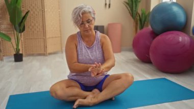 Gri saçlı yaşlı bir kadın spor salonundaki yoga minderinde bacak bacak üstüne atmış acı dolu bir ifadeyle bileğini tutuyor.