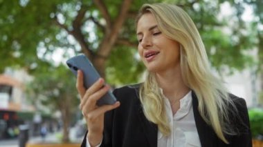 Şehir parkında akıllı telefon tutan bir kadın açık havada yemyeşil alanlar arasında güvenli bir şekilde profesyonel kıyafetlerle iletişim kurarak modern teknoloji ve bağlantıyı öneriyor.