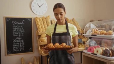 Çizgili önlüklü genç kadın elinde kruvasan tepsisiyle fırında gülümseyen ekmek ve hamur işleri raflarda sergileniyor.