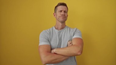 Kendine güveni tam, sarı bir zemine karşı duran, kolları çaprazlanmış, rahat bir şekilde gri bir gömlek giyen, kendine güvenini ve pozitifliğini ifade eden, kendinden emin bir adam.