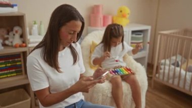 Bir kadın ve kız yatak odasında, kadın telefon kullanırken kız ksilofon çalıyor, oyuncaklar ve bebek mobilyalarıyla çevrili, aile sıcaklığını ve içeride etkileşimi ifade ediyor..