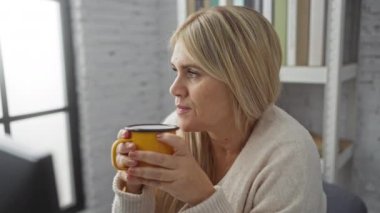 Parlak ofiste kahve içen kadın, pencere kenarında kitap rafları ve bilgisayar ile profesyonel bir ortamda rahatlama ve rahatlama hissi sergiliyor..