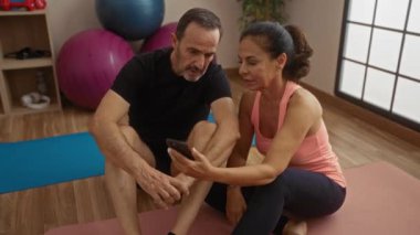Orta yaşlı İspanyol çift, yoga minderleri üzerinde spor salonunda oturuyorlar. Erkek ve kadın, yakınlarda spor toplarıyla akıllı telefona bakıyorlar. Spor ortamında spor yapma ve iletişim kurma yeteneğine işaret ediyorlar.
