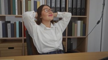 Arka planda kitap ve klasörlerle dolu bir ofiste dinlenen genç bir kadın.
