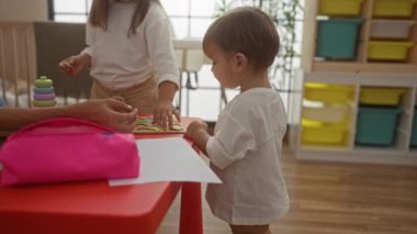 Kadın, oğlu ve kızıyla birlikte parlak bir anaokulunda, masada eğitici oyuncaklarla oynuyor. Aile bağlarını ve erken çocukluk gelişimini teşvik ediyor..