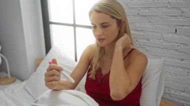 Kırmızı gecelikli bir kadın, modern bir yatak odasındaki bir yatakta oturmuş hamilelik testine bakıyor. Güneş ışığı pencereden süzülürken endişeli bir görünüm sergiliyor..