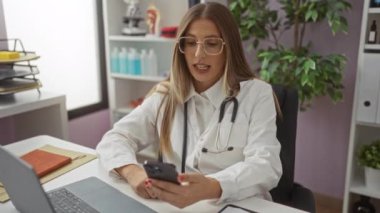 Klinikteki kadın doktor, gözlük ve steteskop takıyor. Bilgisayarlı ve klasörlü akıllı telefon kullanıyor. Klinik bir ortamda ciddi bir video görüşmesine dalmış..