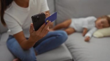 Gri bir kanepede oturan bir kadın, elinde telefon ve kredi kartıyla, yanında uyuyan bir bebekle oturmuş, teknolojisiyle sıcak bir aile anını sergiliyor..