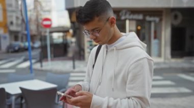 Şehir sokağında gözlüklü ve beyaz kapüşonlu akıllı telefon kullanan genç adam.