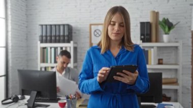 Mavili kadın, modern ofiste masa başında çalışan bir adamla tablet kullanıyor. Takım çalışmasını ve teknolojisini vurguluyor.