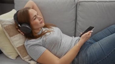 Modern bir oturma odasında bir kanepede dinlenen, kulaklıkla müzik dinleyen, akıllı bir telefon tutan, çağdaş bir evde huzurlu bir an yaşatan bir kadın..