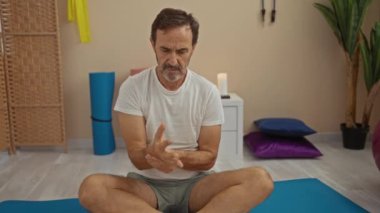 Orta yaşlı bir adam spa odasında bacak bacak üstüne atarak oturuyor. Sakin bir ifadeyle bileğini tutuyor. Yastıklarla ve barışçıl dekorlarla çevrili..