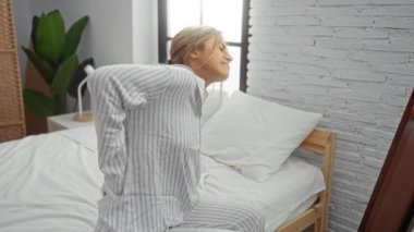 Modern bir yatak odasında beyaz bir yatakta sırt ağrısı çeken, çizgili pijama giyen bir kadın, evde rahat bir sabah manzarası olduğunu düşündürüyor..