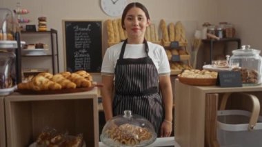 Fırında çalışan genç bir kadın. Etrafı ekmek, pasta ve glütensiz kurabiyelerle çevrili. Çizgili bir önlük takıyor..
