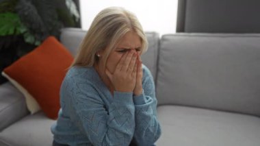 Modern bir oturma odasındaki kanepede ağlayan bir kadın, ellerini yüzünü örten, mavi bir kazak giyen, arka planda turuncu yastıklı ve çiçekli bir kadın..