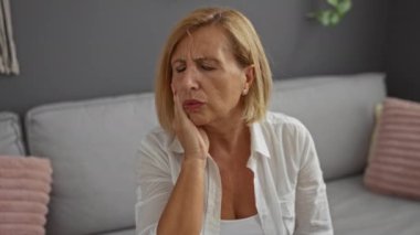 Oturma odasında beyaz gömlekli, kısa sarı saçlı, kanepe ve yastıklı bir kadın diş ağrısı çekiyor.