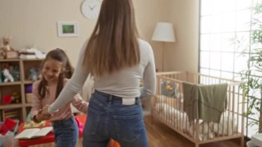Anne ve kızı, sevimli bir oyun odasında, oyuncaklarla ve beşikte neşeyle dans ediyor. İçeride sevgi dolu bir aile bağının altını çiziyor..
