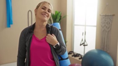 Kadın spor salonunda yoga minderini tutarken gülümsüyor pembe tişört giyiyor ve spor aletleri arka planda fitness aktivitesi için görünen parlak iç mekan ayarlarıyla ukalalık yapıyor..