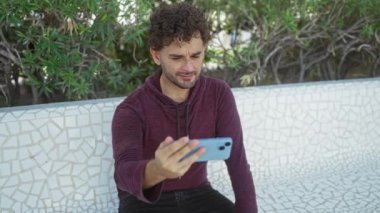 Akıllı telefonlu bir adam parkta oturmuş gülümsüyor ve kentteki yaşam tarzını gözler önüne seren yapraklı arka planda günlük giysiler içinde selfie çekiyor.