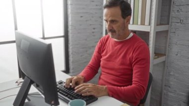 Ofiste kırmızı süveter giyen orta yaşlı İspanyol bir adam, bilgisayarla masada oturuyor, raflar ve kitaplarla iç mekanda endişeli görünüyor..