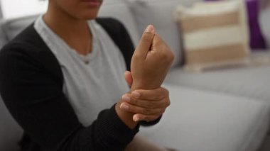 Oturma odasında bileğini tutan, kıvırcık saçlı ve odaklanmış ifadeli bir kadın evde acı çektiğini gösteriyor..